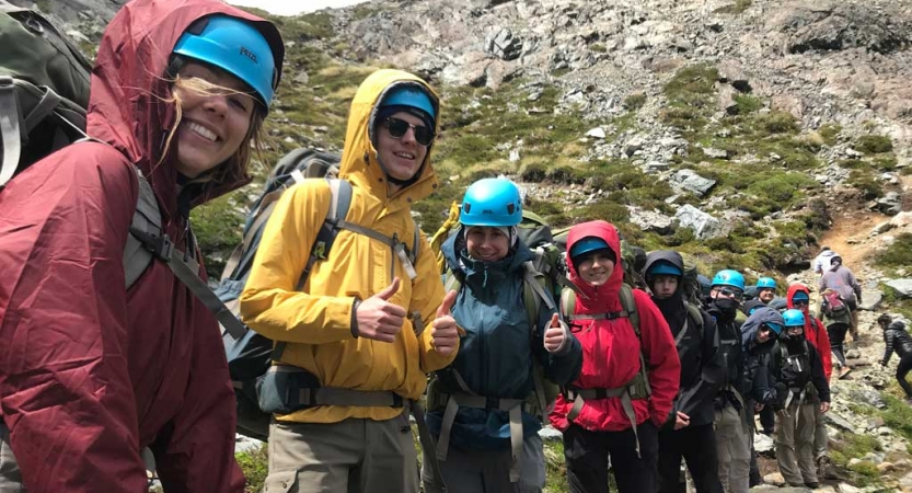 A group of people wearing safety gear and backpacks smile at the camera while hiking along a rocky trail.
